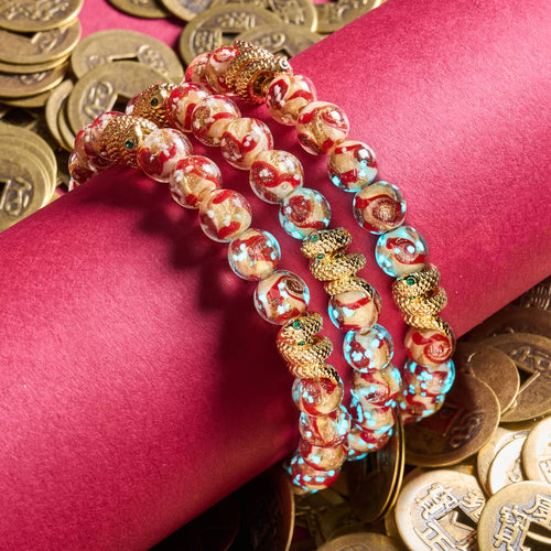 Golden Snake Firefly Glass Swirl Bracelets with glowing red and gold beads and 18k gold-plated snake charms on red fabric with gold coins by NOGU